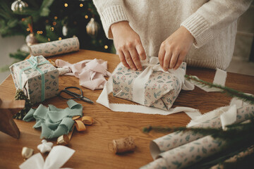 Woman wrapping christmas gift with stylish ribbon on wooden table with festive decorations in...