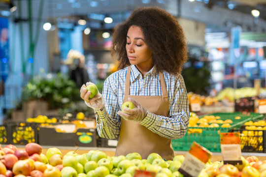Serious concentrated woman seller shop worker inspects sorts and sorts apples in supermarket, among fruits and vegetables in grocery department.