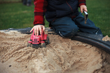 Close up photography of a boy playing with his plastic red tractor in an outdoor sandpit