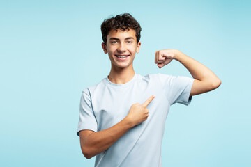 Handsome strong boy wearing dental braces, pointing finger at biceps, looking at camera