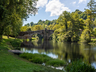 Vista del puente romano sobre el río tranquilo en Allaritz, rodeado de árboles verdes cielo azul y nubes blancas, en Orense, viajando por Galicia España en verano de 2021 - obrazy, fototapety, plakaty