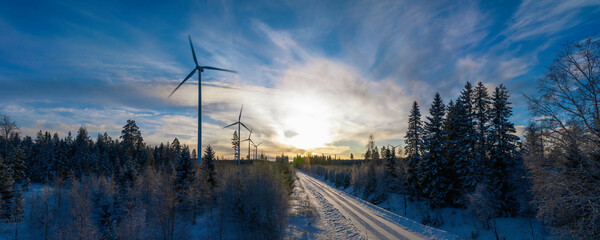 Panoramic wide scenic aerial photo over forest winter road with windmills standing in row at the left side in forest. Road at the right side of view. Large wind turbines with blades