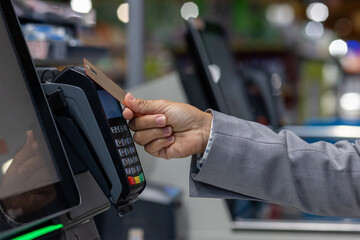 Close-up photo of the hand of a female buyer paying in a supermarket store with a bank credit card at the self-service checkout. - obrazy, fototapety, plakaty