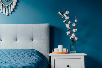 Stylish modern bedroom in dark colors. Cozy interior with navy blue walls, home decor. Bed with gray fabric headboard, blue blanket, bedside table, vase with natural cotton flowers, candles on tray.