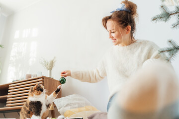 Cat pet is playing with glass bubble during young woman decorating Christmas tree with her cat in light modern Scandinavian interior. Winter festive holidays with domestic pet at home. Selective focus