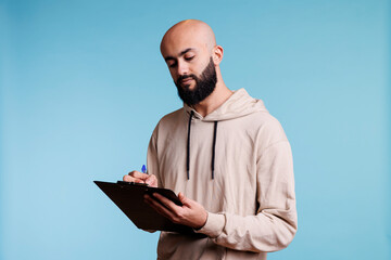 Young arab man writing in clipboard with concentrated facial expression. Focused arabian bald bearded person making list and taking notes while standing on blue background