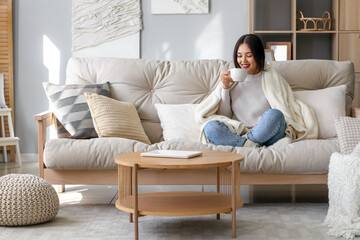 Young Asian woman with warm plaid drinking tea at home - obrazy, fototapety, plakaty