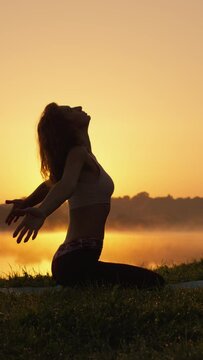 Nature as a source of inspiration for physical and spiritual development. Bright sunset sunrise over lake as background for yoga practice and meditation in vertical video silhouette of slender woman.