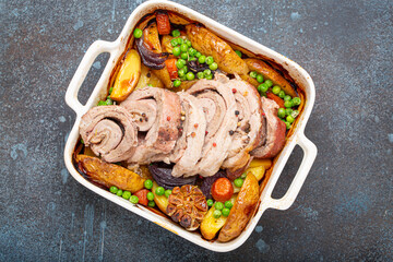 Rolled sliced pork roasted in white casserole dish with potatoes, vegetables and herbs on blue dark...