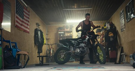 A male mechanic rolls out his motorcycle from his workshop garage with numerous tools and a US flag. Completion of repairs and test drive of the new bike