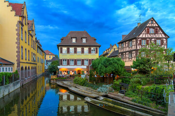 Twilight evening views of half timbered buildings and illuminated waterfront cafes on the Lauch River in the historic medieval Little Venice district of Colmar, France, in the Alsace region.	