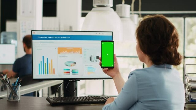 Employee looks at greenscreen on phone, working on business operations at desk. Person holding smartphone device with isolated display showing mockup template, blank layout screen.
