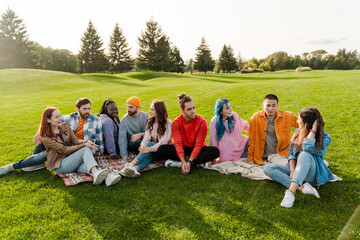 Group of smiling attractive multiracial friends wearing colorful clothing meeting, talking, relaxing, chilling, sitting on green grass in park. Diversity, friendship, travel concept  - obrazy, fototapety, plakaty