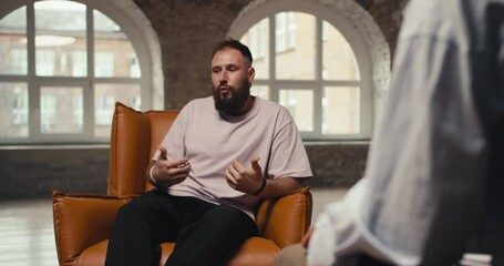 An excited man with a beard in a pink shirt talks about his problems and questions to a psychologist. A young man sits on a brown chair and communicates with a psychologist in a special room made of