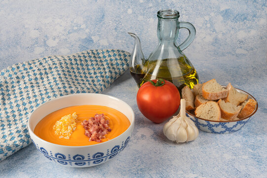 Still life of a bold or bowl with salmorejo and its ingredients, bread, garlic, oil and tomato isolated on a light blue marbled background