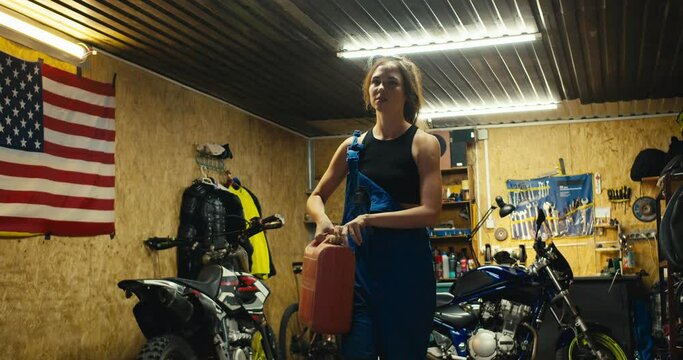 A tired girl mechanic in a black top and blue overalls comes out of her garage workshop along with a canister of water and pours herself near the entrance to the garage. An impromptu shower after a