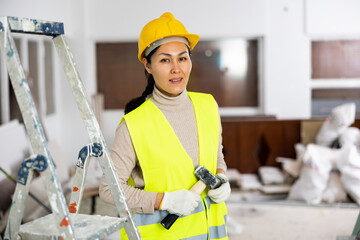 Portrait of positive Asian woman builder with hammer standing in apartment.
