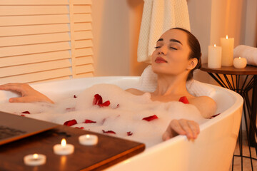 Woman with laptop taking bath in tub with foam and rose petals indoors