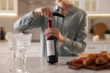 Romantic dinner. Woman opening wine bottle with corkscrew at table in kitchen, closeup