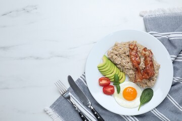 Delicious boiled oatmeal with fried egg, bacon, avocado and tomato served on white marble table, flat lay. Space for text
