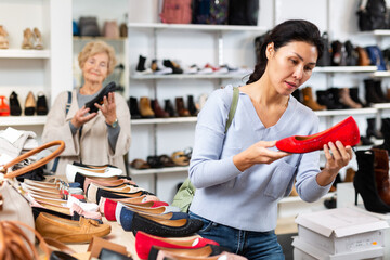 Asian woman selecting new shoews while standing in salesroom shoeshop. Elderly European woman shopping in background.