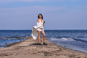 Fototapeta na wymiar Portrait of attractive brunette girl with long hair posing on a deserted beach. She wears white shorts, a top, a long white cape. She is looking to the camera.
