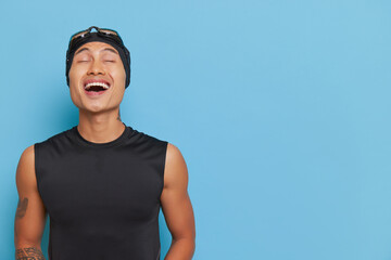Young asian boy wearing black sleeveless top and swimming cap with goggles laughing over blue...