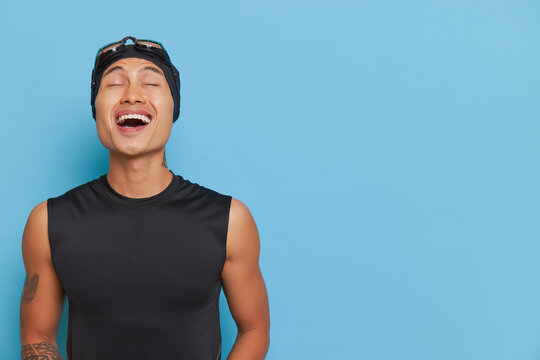 Young asian boy wearing black sleeveless top and swimming cap with goggles laughing over blue backdrop, sport life concept, copy space