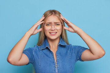 Pretty blonde in blue denim dress pressing hands to her head standing on blue background, bad day concept, copy space