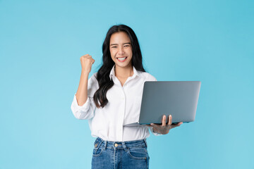 Cheerful beautiful Asian woman holding laptop with fists clenched celebrating victory expressing...