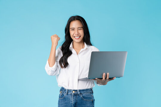 Cheerful beautiful Asian woman holding laptop with fists clenched celebrating victory expressing success on blue background.