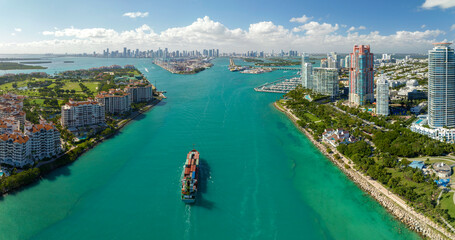 Aerial view of large container ship entering in Miami harbor main channel near South Beach. Luxurious hotels and apartment buildings on waterfront and high skyscrapers of downtown district in distance