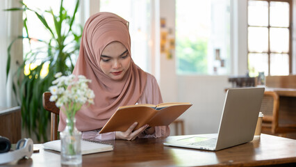 A concentrated young Asian-Muslim woman is reading a book while working remotely at a coffee shop.