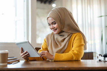 An attractive and happy Asian-Muslim woman is using a digital tablet while relaxing in a coffee shop