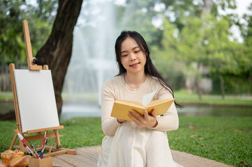 A beautiful Asian woman enjoys reading a book while picnicking in a green park on the weekend alone.
