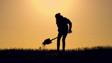 Woman digging pit with shovel dark silhouette in farm field at sunset time