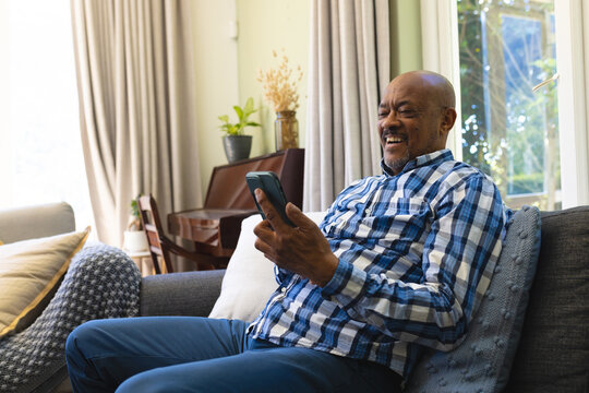 Happy african american senior man sitting on sofa, using smartphone and smiling in sunny living room