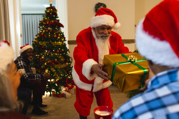 Happy biracial man in santa clothes giving gift to friend next to christmas tree at home