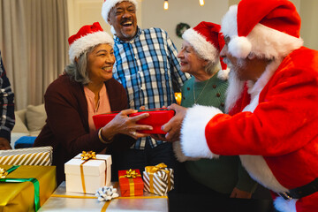 Happy biracial man in santa clothes giving gifts to friends in living room at home