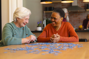 Happy diverse senior female friends playing with jigsaw puzzles in sunny dining room at home