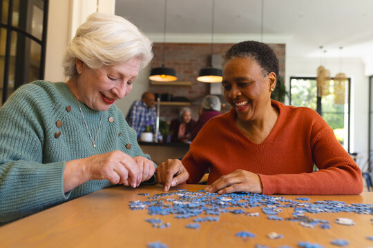 Happy diverse senior female friends playing with jigsaw puzzles in sunny dining room at home