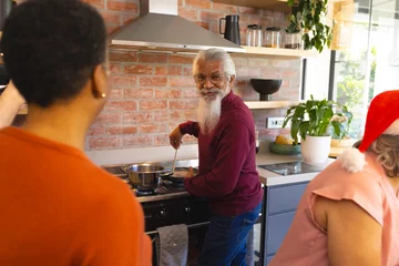 Rolgordijnen Happy diverse group of senior friends in santa hats, preparing meal in sunny kitchen at home © wavebreak3