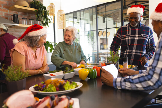 Happy diverse group of senior friends in santa hats, preparing meal in sunny kitchen at home