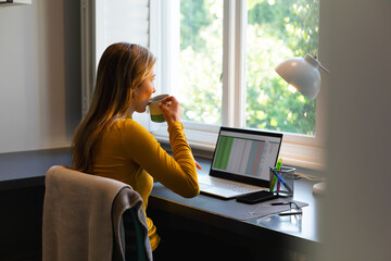 Caucasian woman sitting at desk, working from home with laptop and drinking coffee in sunny room