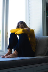 Sad caucasian woman sitting on windowsill and hiding face in sunny room at home