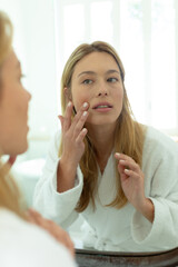 Happy caucasian woman in bathrobe looking in mirror and touching face in sunny bathroom