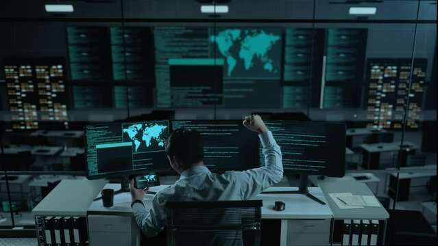 Back View Of Happy Asian Man Developer Looking At Code On Mobile Phone While Write Code With Multiple Computer Screens In The Office
