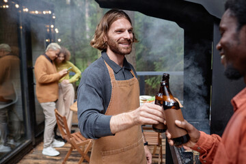 Happy young men drinking beer at party outdoors while they frying meat