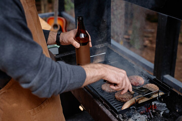 Close-up of young man drinking beer and frying meat on grill outdoors