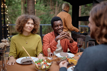 Group of young friends talking to each other while sitting at table and eating meat at party outdoors